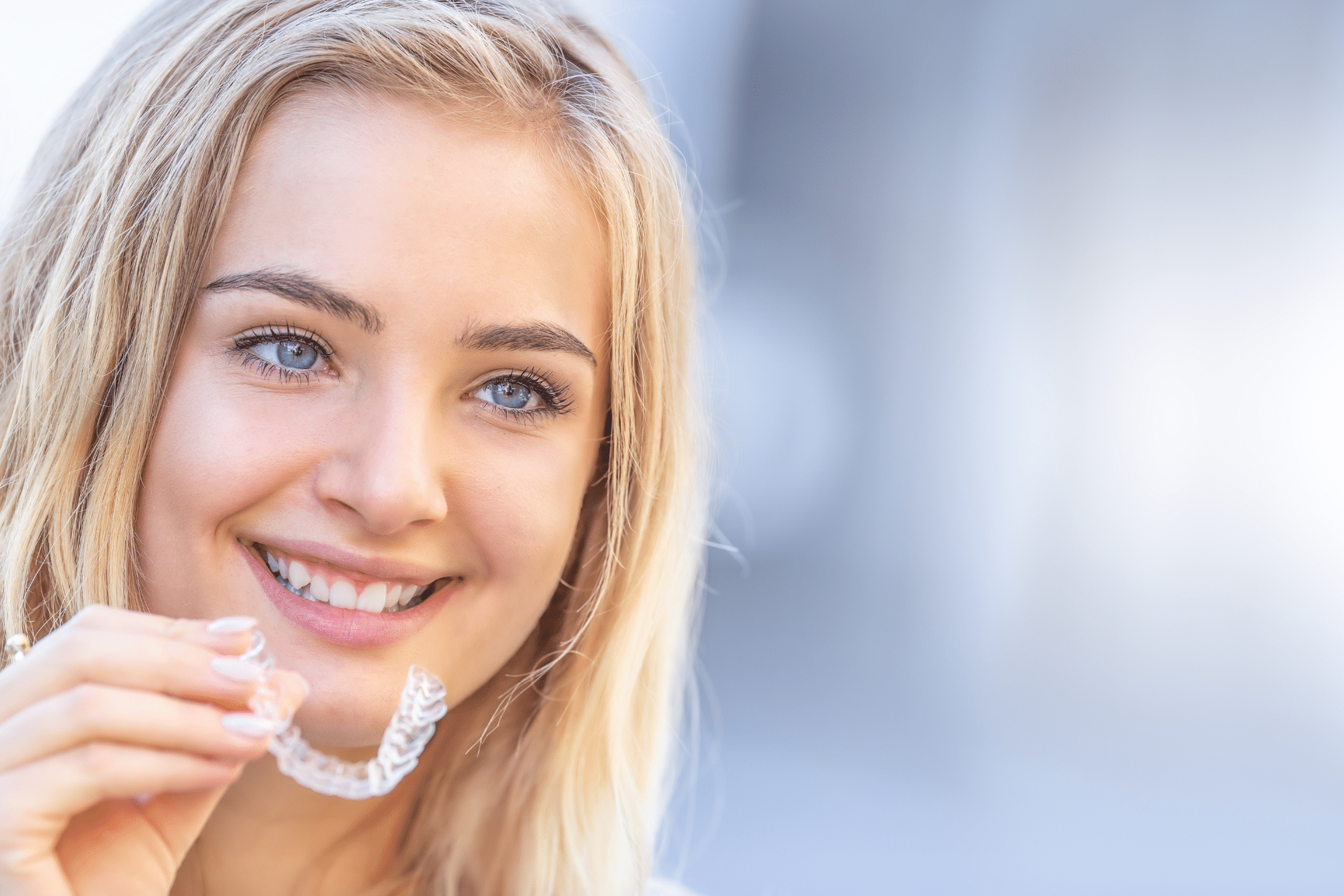 A smiling blonde woman holding a clear aligner, a treatment for straighter smiles available in Rockford & Winnebago, IL.