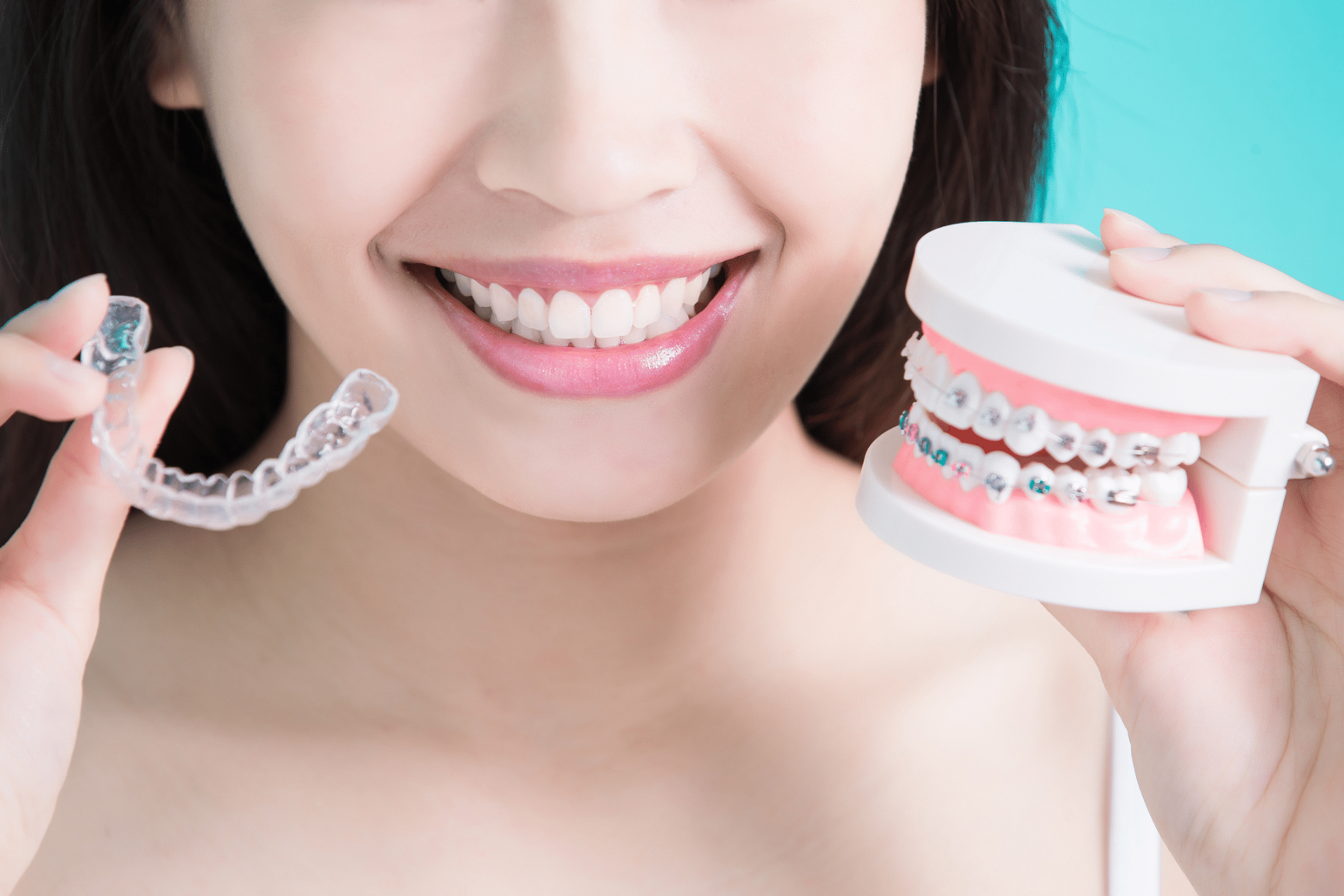 A close-up of a smiling woman holding both a clear aligner and a model of metal braces.